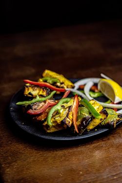 Grilled paneer cubes served on a black plate with sliced green and red bell peppers, onion rings, and a lemon wedge.