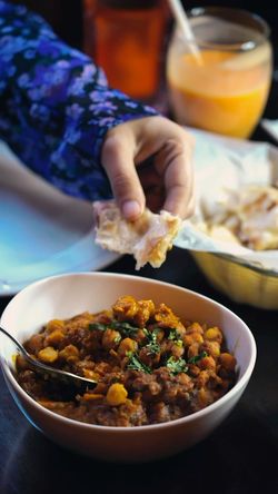 Hand dipping garlic naan into a bowl of chickpea curry garnished with herbs, with a drink and additional naan in the basket.
