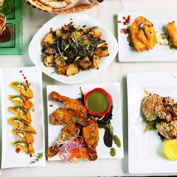Variety of Indian dishes including tandoori chicken, fried appetizers, and spiced potatoes served on white plates.
