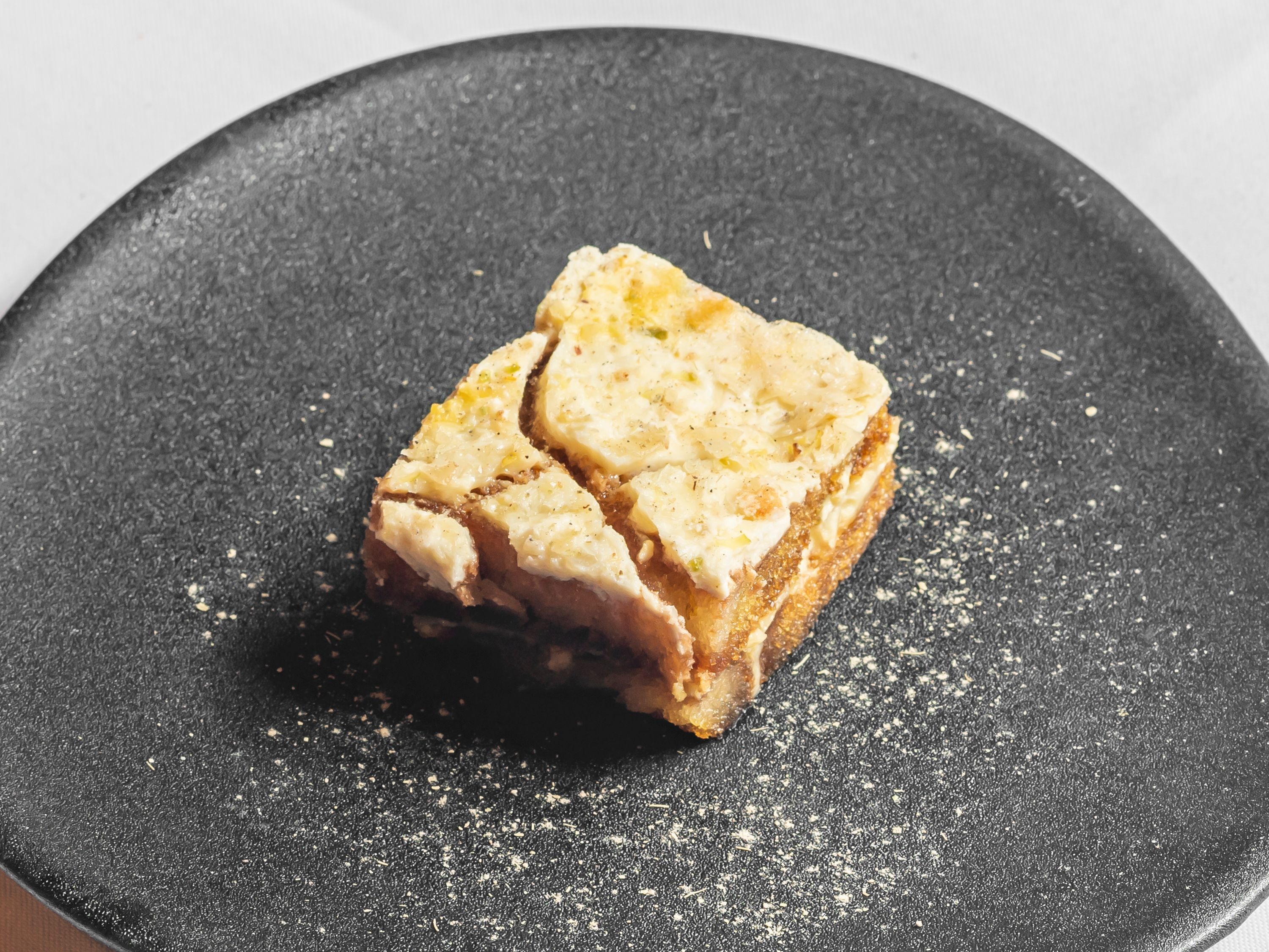 A square piece of Indian dessert, likely a barfi, served on a dark plate with powdered garnish around it.